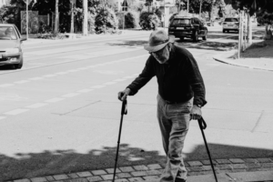 An elderly man walks along a pavement holding two walking canes for support.