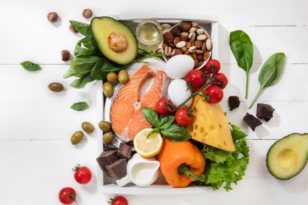 healthy foods including salmon, olives, avocados, spinach, and eggs on a white table