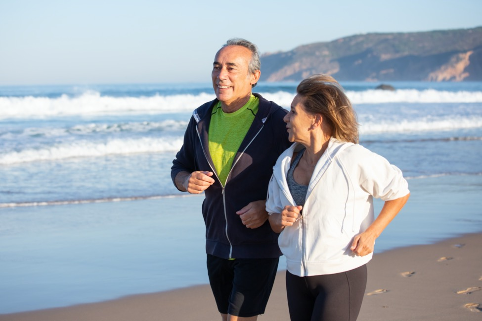An older man and woman jog along a beach