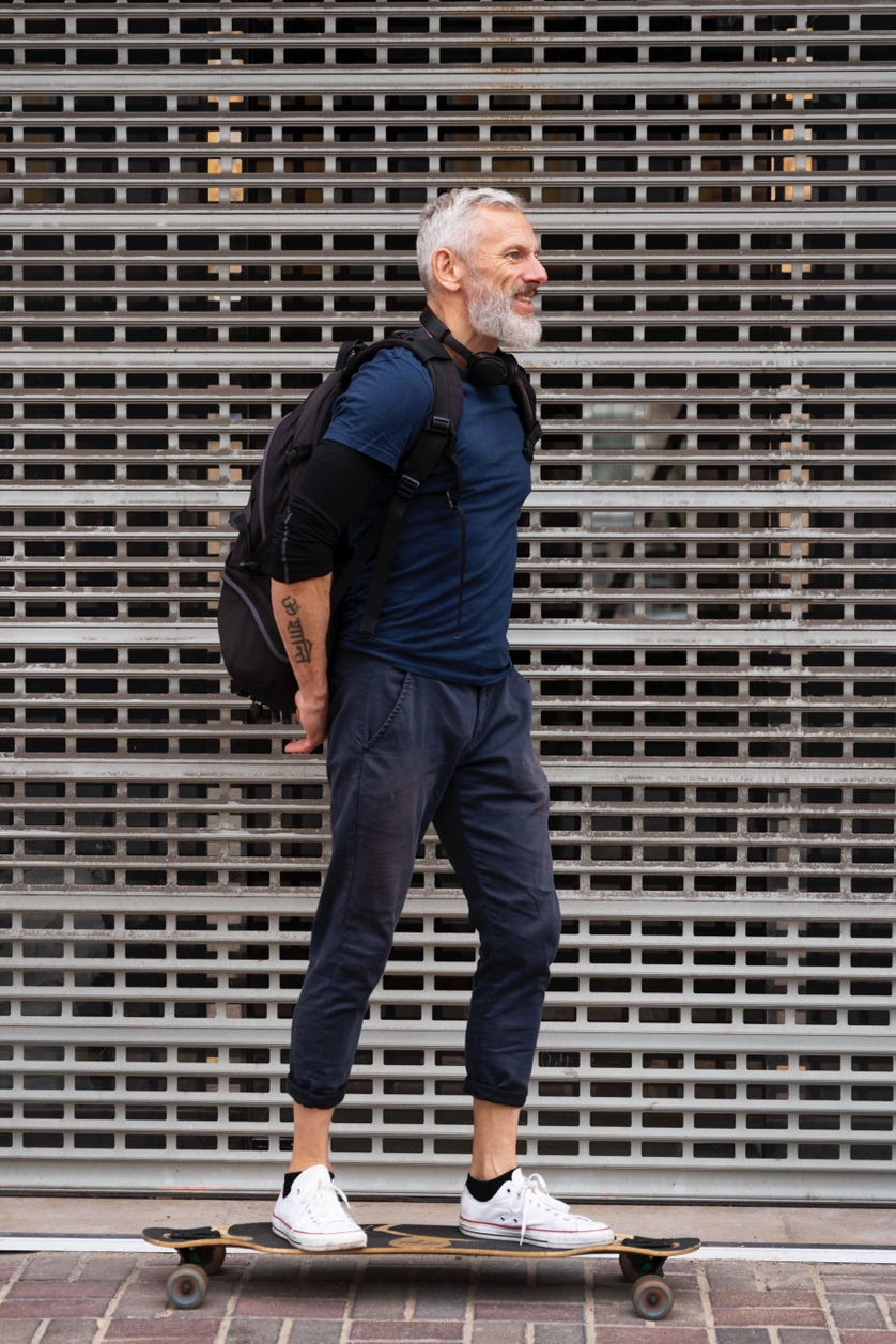 A man with a gray beard and backpack rides a skateboard in front of a closed retail store.