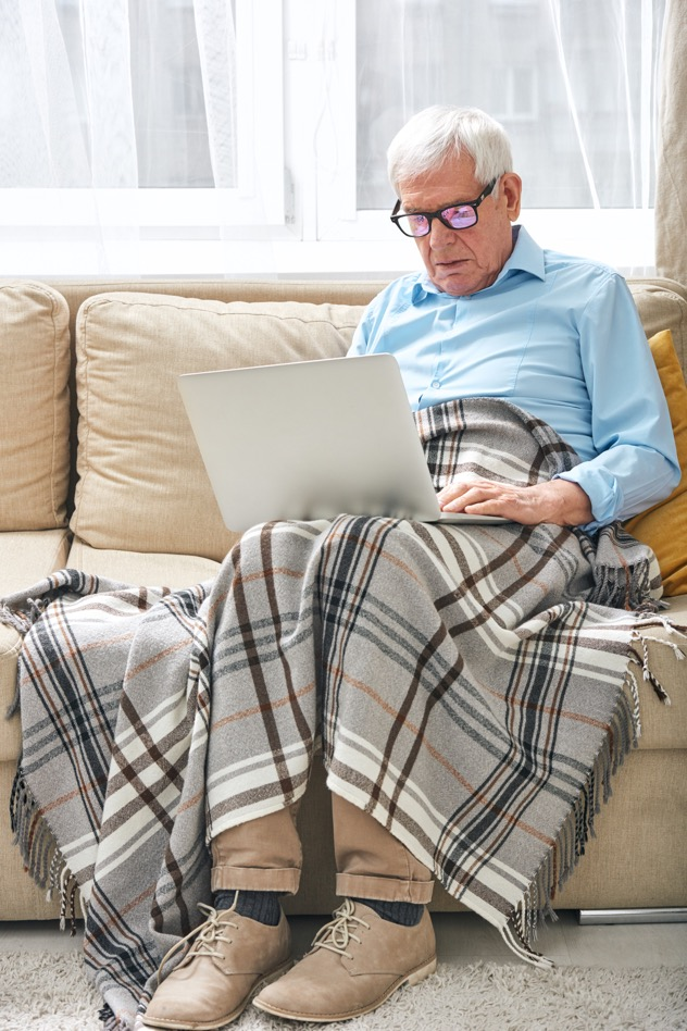 An older man sits on a sofa with his laptop and throw blanket on his lap. 