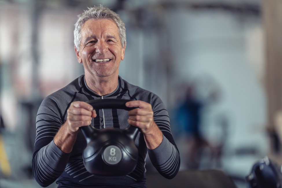 An older man smiles while exercising with a kettlebell.