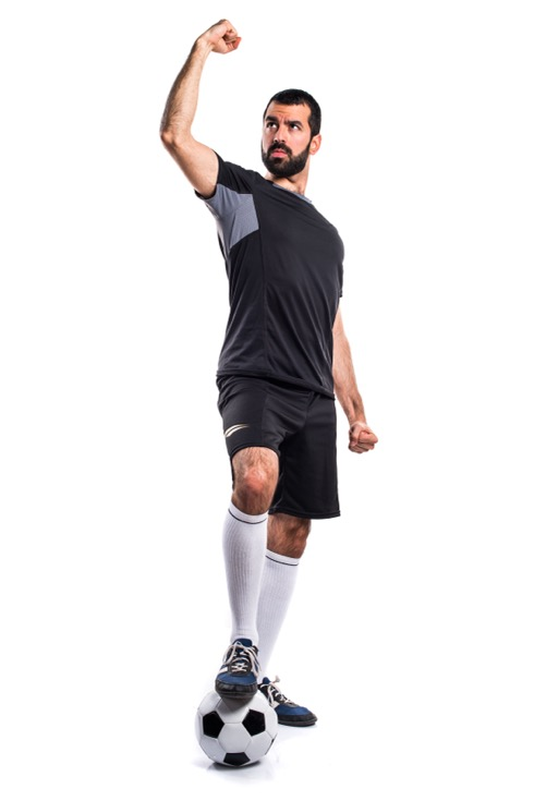 A male soccer player with a beard stands with his foot on the ball while holding his fist in victory.