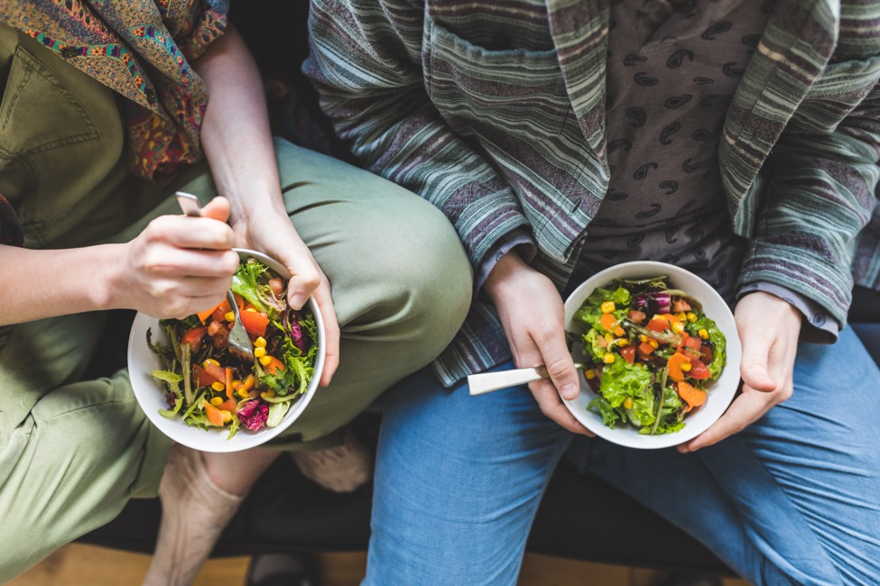 A couple sitting together holding bowls of salad. 